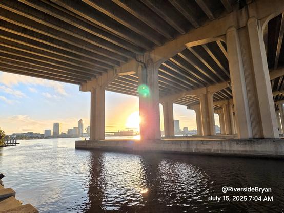 Standing beneath a massive highway overpass bridge at a river's edge overlooking the downtown skyline where a bright morning sun is rising in thr bright sunny sky.