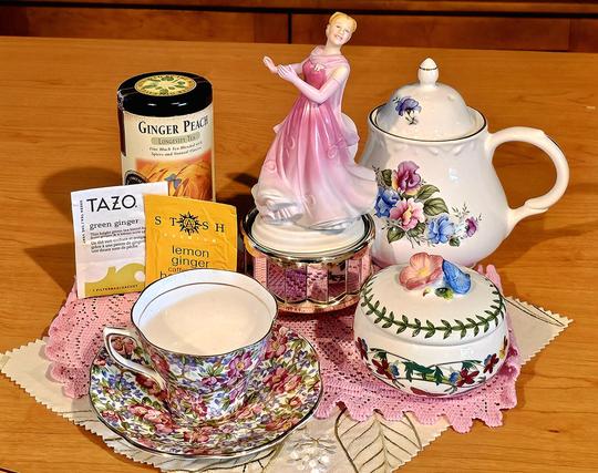 Tea tableau celebrating Ginger Rogers, centered around a porcelain figurine of her dancing in a pink ball gown on a mirrored pedestal. There is floral china, a pink crochet doily, an ivory embroidered mat, and three different ginger- flavored teas.