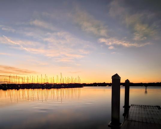From a darkened, shadowy shoreline and pier overlooking a calm waterway with a large marina of sailboats docked in silhouette against a colorful sunrise climbing above the horizon and casting reflections across the sky and onto the water's surface below.