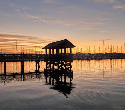 From a darkened, shadowy shoreline and pier overlooking a calm waterway with a large marina of sailboats docked in silhouette against a colorful sunrise climbing above the horizon and casting reflections across the sky and onto the water's surface below.