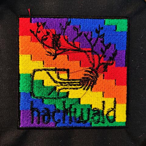 Ein Foto des gestickten hackwald-Logos über einer stufigen Regenbogenflagge auf einer schwarzen Patchbasis. Das hackwald-Logo ist ein Chaosknoten, aus dem die Kabel nach oben herauswachsen wie Äste. Es gibt Blätter, eine hängende Fledermaus und eine sitzende Eule zwischen den Zweigen.