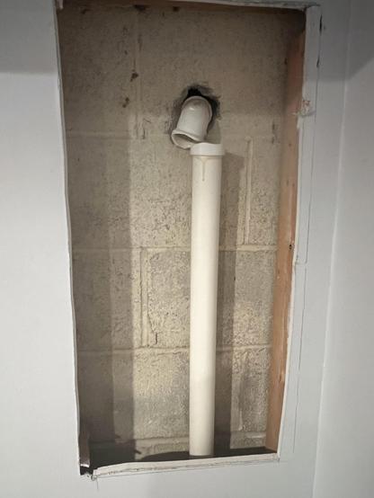 An open wall section showing a rectangular opening. Inside, there is a white plastic pipe extending vertically, disconnected from a fitting that emerges from the exposed concrete wall behind it.