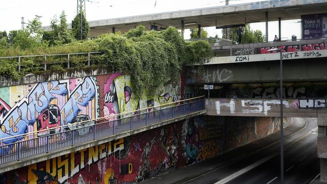 urban scene of a graffitied concrete bridge across a highway with a station on top, also lots of greenery