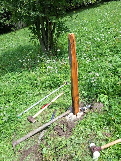 A freshly installed rustic wooden fence post stands upright in a grassy area surrounded by wildflowers. The post is natural, unevenly shaped, and appears to be made from solid wood, with some charring visible. It is secured in the ground with a ring of stones. Various hand tools are scattered around on the grass, including a wooden-handled pickaxe, a sledgehammer, a rubber mallet, pliers, and asprinkler with a metal bar. In the background, there is a bushy tree and a lush green meadow.