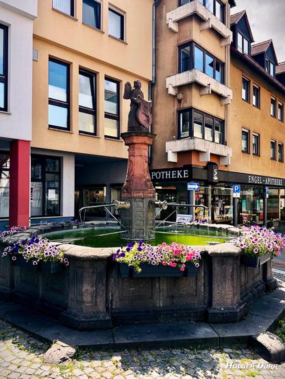 Stimmungsvolle Aufnahme des Engelsbrunnens aus leicht seitlicher Perspektive. Der Blick öffnet sich zum Platz mit umliegenden Gebäuden, darunter eine Apotheke. Der Brunnen mit dem Engel und den blühenden Blumen bildet das ruhige Zentrum des sommerlichen Stadtbildes.