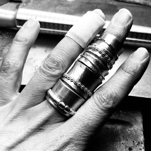 A close-up of a hand adorned with multiple silver rings. One finger has a bandage, indicating a minor injury. The background shows a work surface with tools, suggesting a jewelry-making context. The image is in black and white.