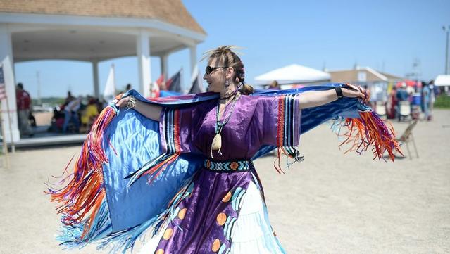 A woman wearing colorful clothing, including a shawl, dancing on a beach. In the distance is a gazebo and a sailboat in the ocean.
