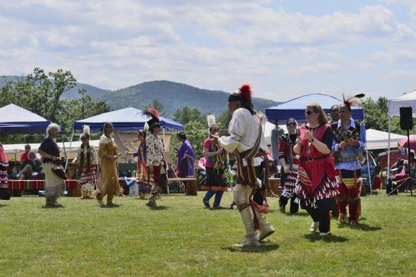 People dancing in circle at a Pow Wow. They are wearing colorful clothing, and dancing on a grassy area. In the distance are mountains and clouds.