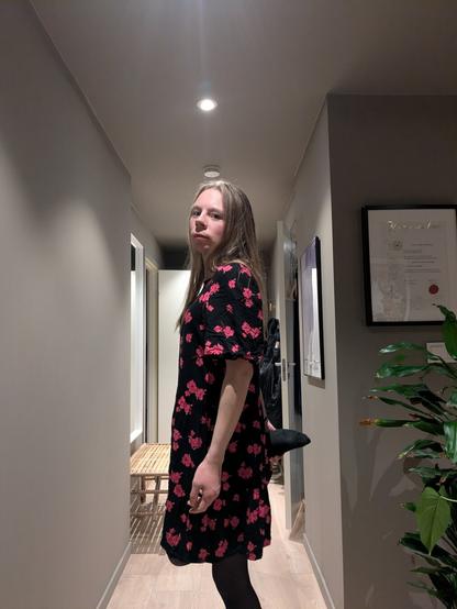 Gender-fluid person stands in a hallway wearing a black floral dress with pink flowers, holding her feet. The setting is warmly lit and cozy, with plants and framed art.