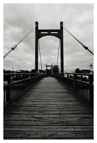 Eine Hängebrücke aus Holz mit Stahlseilen führt perspektivisch ins Bild hinein. Der Himmel ist wolkenverhangen, die Aufnahme ist in Schwarz-Weiß gehalten. Im Hintergrund sind Bäume, Gebäude und ein Kirchturm zu erkennen.