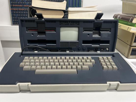 An Osborne 1 portable computer with a small screen and built-in keyboard is open, showing various ports and drive slots. It is placed on a table with several stacked books in the background.