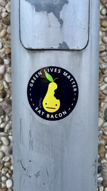 Sticker on an electric pole representing a pear on a dark background. The text around the pear reads « GREEN LIVES MATTER - EAT BACON »