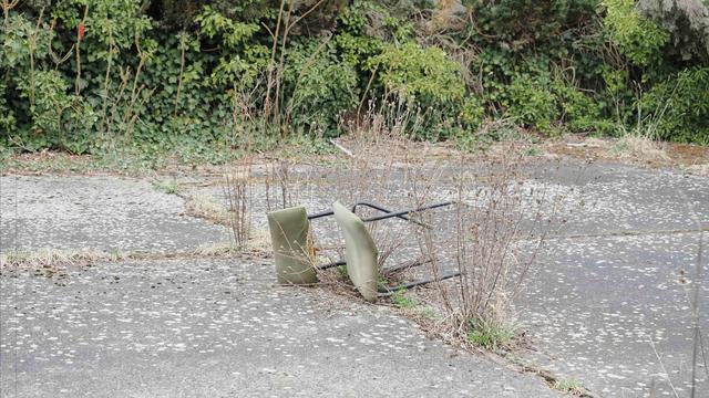 A green metal chair lies overturned on a cracked, overgrown concrete surface. Weeds and dry grass sprout around it, and dense green shrubs and foliage cover the background, indicating neglect and disrepair in the area.