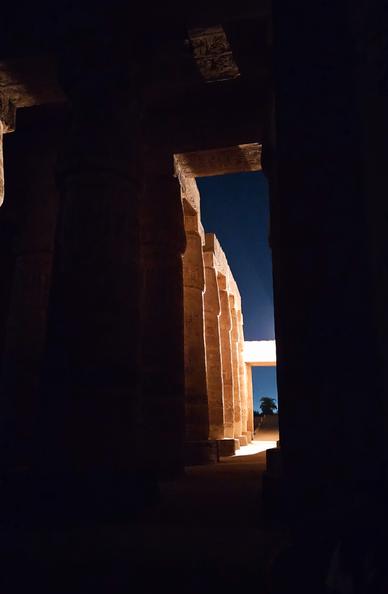 The photo was shot inside the Karnak temple during the light show at night. Most of the surroundings are dark with only slight hints of the temple around us. We're looking along a narrow path through the giant columns standing closely together. They get brighter the further out we look, illuminated by a strong spotlight out of frame. The last column and a stone beam glow brightly in the light. In the far back we see a single palm tree barely visible in front of the last remnants of dusk in the sky.