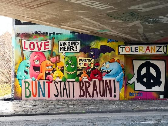 großflächiges, sehr buntes Graffiti:
ein Demozug  aus Phantasiewesen (Monstern?) mit ein bis drei Augen in kräftigen Farben, halten ein Transparent vor dem Körper »bunt statt braun«, auf anderen Schildern steht „love“ und „Bamberg ist bunt“. Ein dreiäugiges grünes Wesen steht zentral und sagt in einer Sprechblase: „Wir sind mehr!“