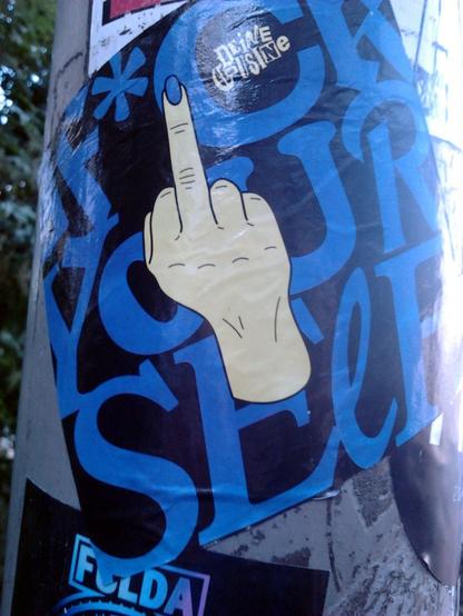 Aufkleber auf einem Laternenmast. Der Text ist blau auf schwarz und wegen dieser Farbwahl und gemischter Schriftarten etwas schwierig lesbar, das Motiv ist weiß und klar. Motiv ist ein ausgestreckter Mittelfinger mit blauem Fingernagel, der den Text teilweise überdeckt. Der Text lautet: Deine Cousine -- F*ck yourself.