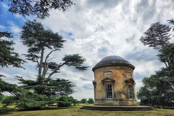 A low-angle shot of a circular, classical stone building with a domed roof and rectangular windows, set against a dramatic sky filled with white and grey clouds. Large, dark green conifer trees frame the building on the left and right, their branches reaching towards the sky. The building stands on a slight rise in a grassy, well-maintained lawn.