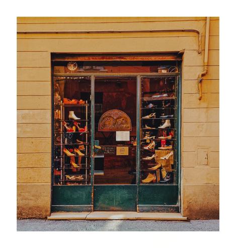 This photo is of the shopfront of a shoe shop called "Mario Calzolaio". The shop front is in a building with a light yellowy beige facade. The shopfront itself features a dark green metal door and big windows with display glass cases filled with various shoes. Small signs and price lists are visible and taped on the inside of the glass door.