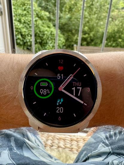 Et smart watch på et håndled. Urkassen er i bronzefarvet metal, og urskiven viser kl. 13:20, dags dato, en puls på 51, skridttæller med 20 skridt og at batteriet er på 98%.