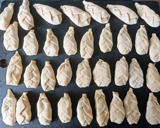 Auf einem Brett ca. 35 fertige Teigtaschen. Sie sind Zapfenförmig und von den Seiten zur Mitte gefaltet, so dass eine schuppige Struktur entsteht.