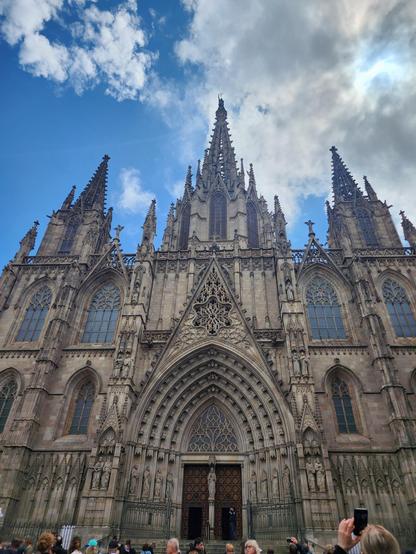 the wide facade of the gothic Barcelona Cathedral, with doors at center and three spires rising into a blue sky.