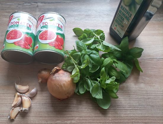 Auf dem Tisch 2 Dosen gehackte Tomaten, viel Basilikum, Zwiebeln, Knoblauch und eine Flasche Olivenöl