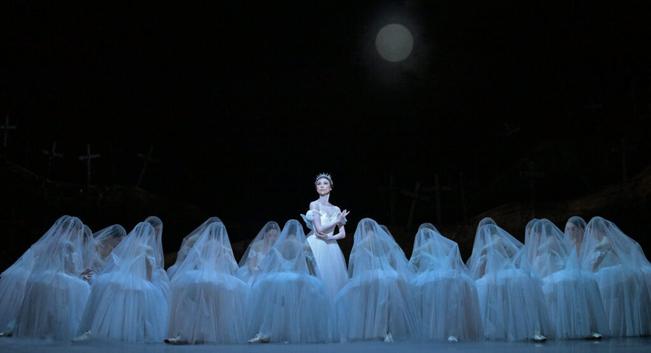 In an eerie scene, a large corps de ballet kneels in lines in gauzy white tutus, with veils draped over their heads. In the middle, in a spotlight, one dancer stands behind them with arms gently folded.
