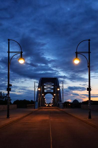 A straight-on, low-angle shot captures the Sternbrücke bridge in Magdeburg at dusk, leading into the distance under a dramatic, cloudy blue sky. Two illuminated, ornate streetlamps with spherical yellow lights frame the foreground. The road surface of the bridge is visible, with a faint white line in the centre. The bridge itself features a series of dark, arched metal trusses with criss-cross patterns. Beyond these, a line of smaller, glowing lights runs along the bridge's path. In the far distance on the right, a dark silhouette of a domed building or structure can be seen against the faint orange glow of the setting sun.