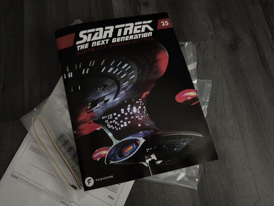 A Star Trek magazine titled "Star Trek: The Next Generation" is displayed, featuring the U.S.S. Enterprise NCC-1701-D on the cover. It is placed on a wooden surface along with plastic packaging and documents.