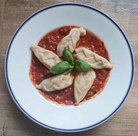 Das fertige Gericht auf dem Teller
5 mit einer schuppenartigen Struktur gefaltete gefüllte Teigtaschen, Tomatensauce und ein Zweiglein Basilikum