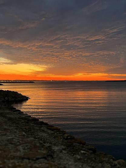 Sundown shot taken from a stone wall: The horizon which appears in fiery red and orange colours is reflected in the dark sea water. Various lights appear on the far coast line.