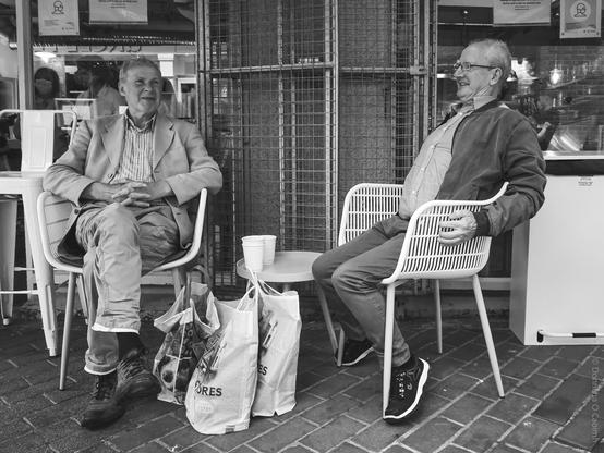 Black and white photograph of two elderly men sitting on white plastic chairs outside a shop, sharing a laugh with shopping bags at their feet and a small table between them.