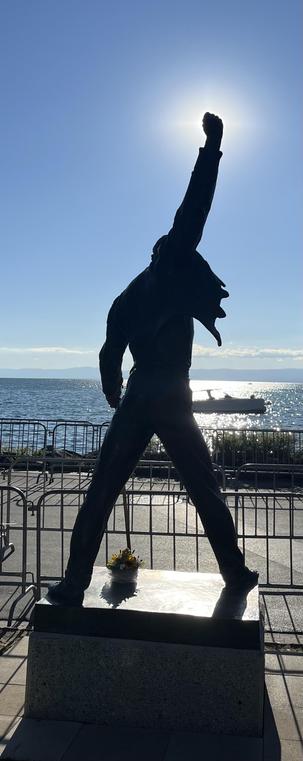 Photo of the Freddy Mercury statue in Montreux, Switzerland from behind. The sky is bright blue, lake Leman is visible, a boat floats on it. Freddy’s right arm characteristically pumps the sky, the fist just covering the sun, looking like it is shining by itself.