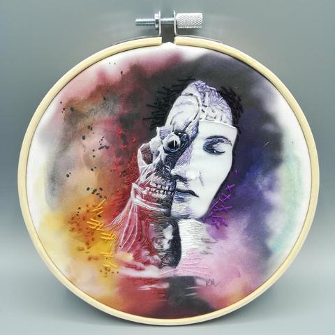 An embroidery hoop holding hand painted cotton with abstract shapes and thread stitches. The white and purple figure depicts a wax teaching model, half face half skull. It is created in tiny stitches of thread.
