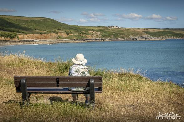 A lady wearing a wide-brimmed hat seated on a bench enjoying the coastal view on a warm summer's afternoon in West Cornwall.