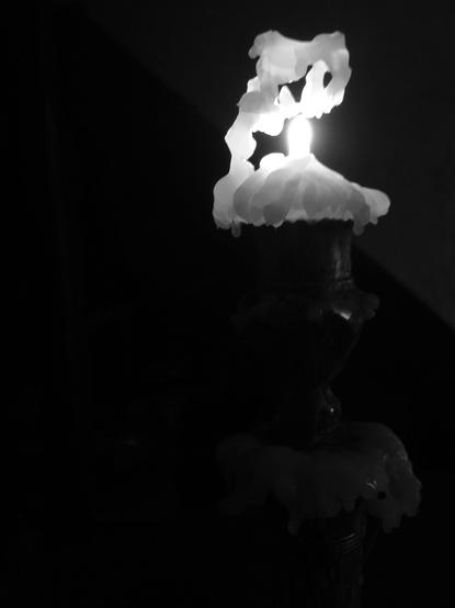 A view of a candle in a metal candlestick, lit only by the flame of the candle itself. The candle has burned down considerably, and the melted wax has formed a kind of gantry above the flame. (Version françæ) Vue d’æn bougie dans æn chandelier métallique, al seulx éclairage de çæt bougie. Eil est très consumæ et læ cire fondux forme æn sorte de portique al-dessus de læ flamme.