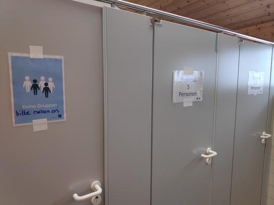 Drei Toilettenkabinen, eine mit Schild "Keine Gruppen, bitte nebenan", zwei mit jeweils "Max. 5 Personen"