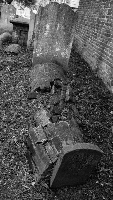 A grave has both a large headstone and a smaller one at the foot of a roughly cylinder shaped (body length) brick construction betweem them. The bricks are broken and part scattered. Other graves are visible behind. Black and white.