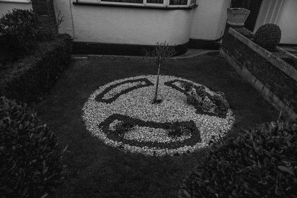 A garden has a circular pebbled area with three shapes in it suggestive of the nuclear symbol. Black and white photo.