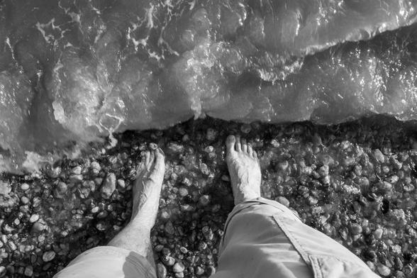 On a pebbled beach, bare (and veiny) feet are about to be hit by an oncoming wave. Black and white photo.