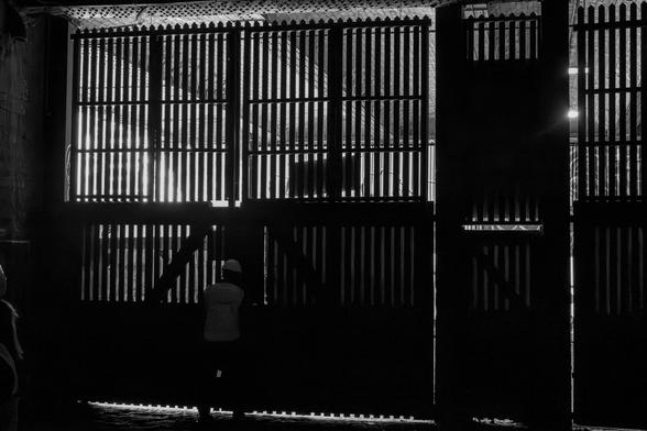 Black and white photo of someone in a hard hat and high vis closing a large slatted gate under a railway bridge.