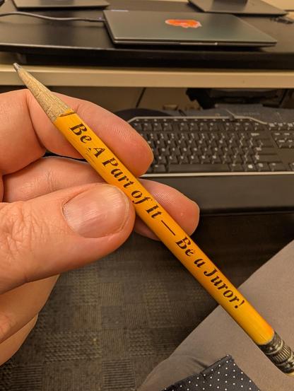The image shows a close-up of a hand holding a yellow pencil. The pencil has the text "Be A Part of It — Be a Juror!" printed in black on its body. The hand is gripping the pencil with the thumb and index finger, and the pencil is positioned diagonally across the image. In the background, there is a desk with a keyboard and a laptop, suggesting an office or study environment. The desk is black, and the keyboard is also black with white lettering. The laptop has a visible sticker on its lid. The hand appears to be resting on a gray fabric surface, possibly a chair or couch. The pencil's eraser is visible at the bottom, and the wood of the pencil is exposed at the tip.