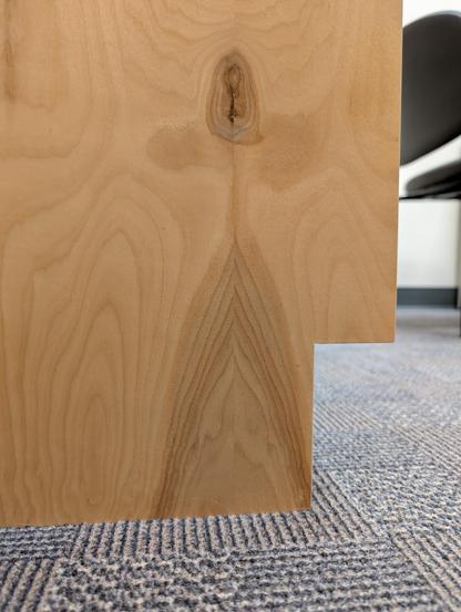 The image shows a close-up view of a wooden surface, likely part of a piece of furniture, such as a table or a desk. The wood has a light, natural color with visible grain patterns, including a prominent knot in the center. The grain lines are smooth and flowing, with a subtle sheen indicating a polished finish. The edge of the wood is cut at a right angle, revealing a clean, sharp line. The background features a patterned carpet with a mix of gray and beige tones, and a portion of a black chair is visible, suggesting an indoor setting. The focus is on the texture and details of the wood, highlighting its natural beauty and craftsmanship.