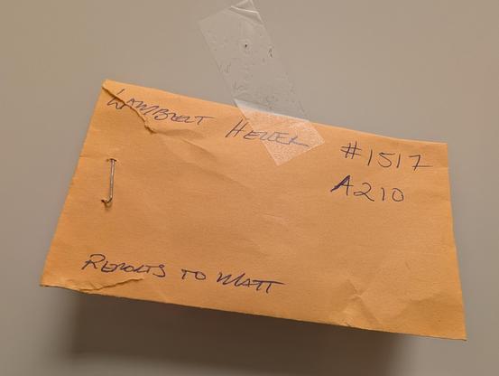 The image shows a brown envelope lying on a flat surface. The envelope is secured with a piece of clear tape at the top and has a metal fastener on the left side. Handwritten text is visible on the envelope. The name "LAMBERT HELLER" is written at the top, followed by the numbers "#1517" and "A210" on the right side. At the bottom, the text "RETURNS TO MATT" is written. The envelope appears to be slightly crumpled, and the handwriting is in blue ink.