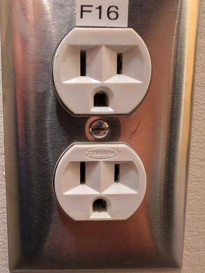 The image shows a close-up of a double electrical outlet mounted on a metallic wall plate. The outlets are white and have the standard two-pronged design with a grounding hole at the bottom. The top outlet is labeled "F16" on a white sticker. The metallic plate is brushed stainless steel, reflecting some light and showing a few minor scratches. The brand "Hubbell" is embossed on the bottom outlet. The wall behind the outlet is a light color, possibly beige or off-white. The outlets are in good condition, with no visible damage or wear.