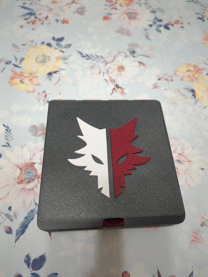 A Gridfinity box, whose outer shell is printed from a dark grey plastic. The box is currently closed, with only the top face visible, which is textured like a print off a textured PEI sheet would be. On the top face is a logo shaped like a wolf, in two halves: one is white, the other is a dark red. The latch is also visible, and it's in the same dark red as half of the wolf.