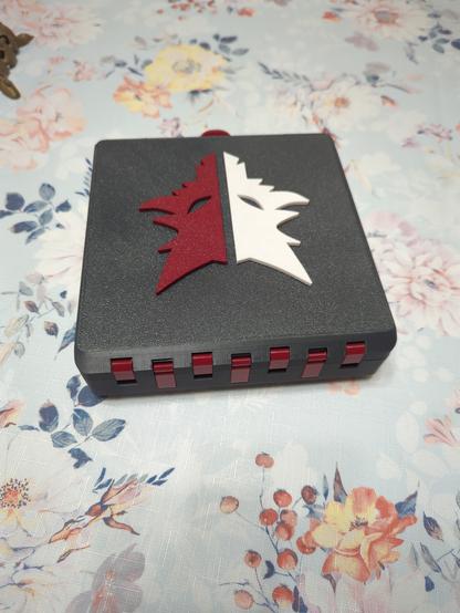 A Gridfinity box, whose outer shell is printed from a dark grey plastic, viewed from the back. The box is currently closed, with only the top face visible, which is textured like a print off a textured PEI sheet would be. On the top face is a logo shaped like a wolf, in two halves: one is white, the other is a dark red. The hinges are also visible, and are printed in the same dark red as half of the wolf.