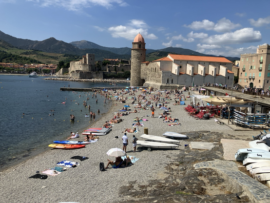Pebble beach with sunbathers, Pyrenees mountains behind stone buildings of old French village