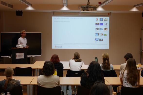 Vortragssituation von hinten: Der Vortragende Alexander Kupfer steht vorne am Pult eines Seminarraums, hält eine Powerpoint-Präsentation und spricht zu seiner Zuhörerinnenschaft.
Bildnachweis: Sommertechnikum MINT / Universität Innsbruck