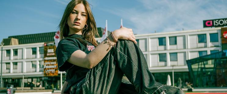 Young woman with long brown hair sits outdoors in a casual pose, wearing a black T-shirt and dark jeans. Behind her, a modern building with large windows and a sign is visible under a blue sky. She is looking directly at the camera with her hands resting on her knees.